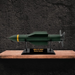 Puede incluir: Un modelo verde oscuro de un misil GBU-57 MOP Bunker Buster, con rayas amarillas y una hélice en el extremo. Se exhibe sobre un soporte negro con texto, en una estantería de madera.
