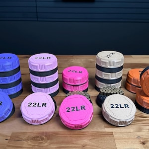 May include: A collection of circular storage containers in blue, light pink, bright pink, beige, and orange. Each lid is embossed with the text 22LR. Some containers are stacked, while others are open, revealing small metallic cylindrical items inside.