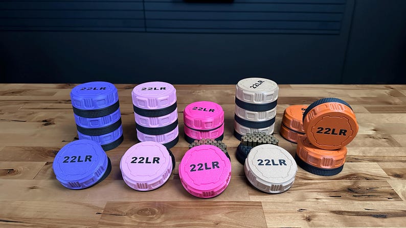 May include: A collection of circular storage containers in blue, light pink, bright pink, beige, and orange. Each lid is embossed with the text 22LR. Some containers are stacked, while others are open, revealing small metallic cylindrical items inside.