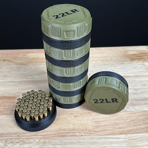 May include: A cylindrical olive green and black storage container with textured grip sections and the text 22LR printed on the top lid. Next to it sits a matching lid and a black base filled with a grid of small brass-colored 22LR cartridges.