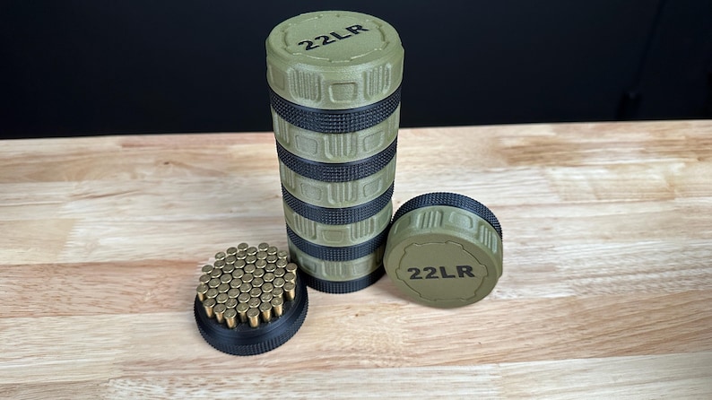 May include: A cylindrical olive green and black storage container with textured grip sections and the text 22LR printed on the top lid. Next to it sits a matching lid and a black base filled with a grid of small brass-colored 22LR cartridges.