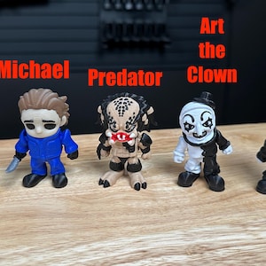 Puede incluir: Cinco peque&ntilde;as figuras de personajes de terror alineadas. De izquierda a derecha: Jason con m&aacute;scara de hockey, Michael con mono azul, Predator, Art the Clown en blanco y negro, y Pinhead con cabeza blanca y traje negro.
