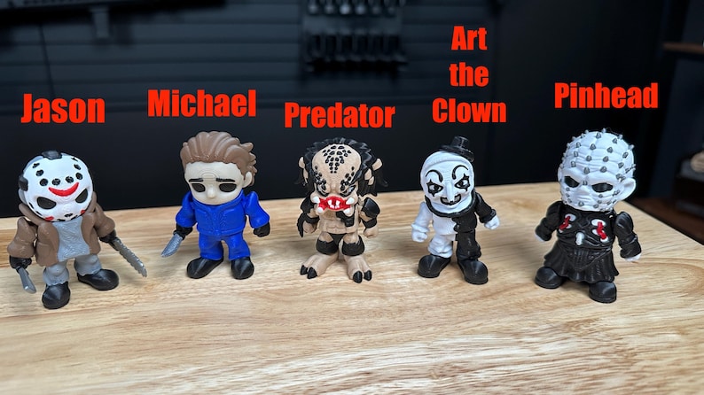 Puede incluir: Cinco peque&ntilde;as figuras de personajes de terror alineadas. De izquierda a derecha: Jason con m&aacute;scara de hockey, Michael con mono azul, Predator, Art the Clown en blanco y negro, y Pinhead con cabeza blanca y traje negro.