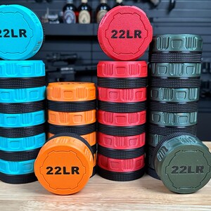 May include: Four cylindrical storage containers in bright blue, orange, red, and olive green, each featuring a black textured grip band and a matching lid embossed with the text 22LR. The containers are stacked in varying heights on a wooden surface.
