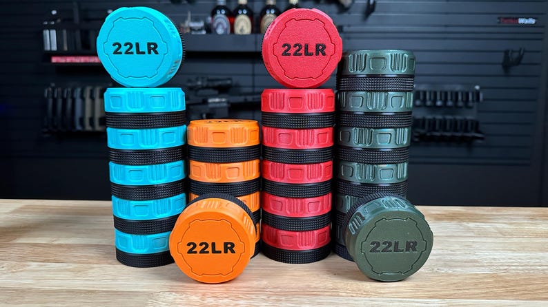 May include: Four cylindrical storage containers in bright blue, orange, red, and olive green, each featuring a black textured grip band and a matching lid embossed with the text 22LR. The containers are stacked in varying heights on a wooden surface.
