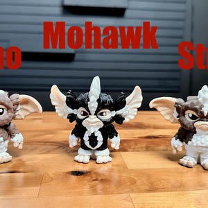 Puede incluir: Tres peque&ntilde;as figuras de personajes Gremlins alineadas. La figura de la izquierda es marr&oacute;n y blanca con la etiqueta Gizmo, la central es negra y blanca con Mohawk, y la de la derecha es marr&oacute;n y blanca con la etiqueta Stripe.