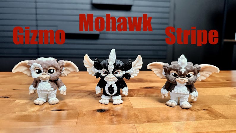 Puede incluir: Tres peque&ntilde;as figuras de personajes Gremlins alineadas. La figura de la izquierda es marr&oacute;n y blanca con la etiqueta Gizmo, la central es negra y blanca con Mohawk, y la de la derecha es marr&oacute;n y blanca con la etiqueta Stripe.
