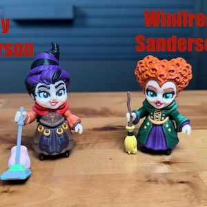 Puede incluir: Dos peque&ntilde;as figuras etiquetadas como Mary Sanderson y Winifred Sanderson. Mary tiene el pelo morado, una bufanda roja y un vestido marr&oacute;n, sosteniendo una aspiradora azul. Winifred tiene el pelo naranja, un abrigo verde y una falda morada, sosteniendo una escoba.