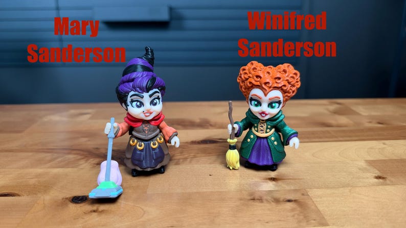 Puede incluir: Dos peque&ntilde;as figuras etiquetadas como Mary Sanderson y Winifred Sanderson. Mary tiene el pelo morado, una bufanda roja y un vestido marr&oacute;n, sosteniendo una aspiradora azul. Winifred tiene el pelo naranja, un abrigo verde y una falda morada, sosteniendo una escoba.