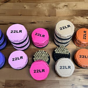May include: A collection of round, plastic ammunition storage containers in purple, pink, tan, and orange. Each lid is embossed with the text 22LR in black. Some containers are stacked, while others are open, revealing rows of small brass cartridges.
