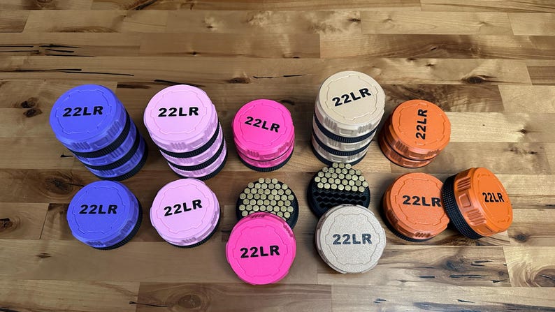 May include: A collection of round, plastic ammunition storage containers in purple, pink, tan, and orange. Each lid is embossed with the text 22LR in black. Some containers are stacked, while others are open, revealing rows of small brass cartridges.