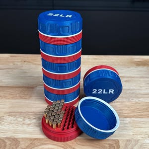 May include: A stack of cylindrical blue and red storage containers with the text 22LR printed on the top lid. One container is open, revealing a red interior tray filled with small brass-colored cartridges, while another lid sits nearby.