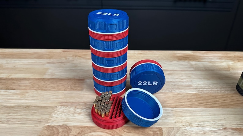 May include: A stack of cylindrical blue and red storage containers with the text 22LR printed on the top lid. One container is open, revealing a red interior tray filled with small brass-colored cartridges, while another lid sits nearby.