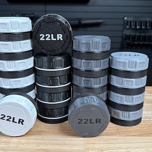 May include: A collection of cylindrical storage containers for 22LR ammunition, arranged in four stacks of varying heights. The containers feature white, black, and gray colors with textured, knurled edges and the text 22LR printed on the lids.