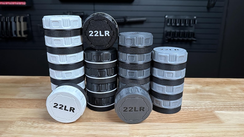 May include: A collection of cylindrical storage containers for 22LR ammunition, arranged in four stacks of varying heights. The containers feature white, black, and gray colors with textured, knurled edges and the text 22LR printed on the lids.