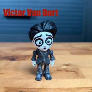 Puede incluir: Una peque&ntilde;a figura de Victor Van Dort con piel blanca p&aacute;lida, ojos grandes y oscuros y cabello negro. El personaje viste una chaqueta de traje gris oscuro sobre un chaleco y pantalones a rayas. El texto Victor Van Dort aparece en rojo.
