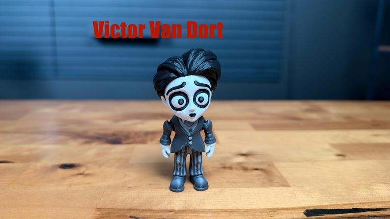 Puede incluir: Una peque&ntilde;a figura de Victor Van Dort con piel blanca p&aacute;lida, ojos grandes y oscuros y cabello negro. El personaje viste una chaqueta de traje gris oscuro sobre un chaleco y pantalones a rayas. El texto Victor Van Dort aparece en rojo.