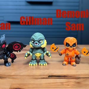 Puede incluir: Cuatro peque&ntilde;as figuras de vinilo dispuestas en fila. De izquierda a derecha: un Mothman negro con ojos rojos, un Gillman verde, un Demonic Sam naranja con una piruleta de calabaza y una figura de Sam naranja con piruleta y saco.