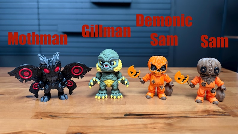 Puede incluir: Cuatro peque&ntilde;as figuras de vinilo dispuestas en fila. De izquierda a derecha: un Mothman negro con ojos rojos, un Gillman verde, un Demonic Sam naranja con una piruleta de calabaza y una figura de Sam naranja con piruleta y saco.