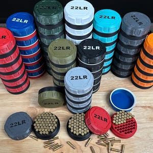 May include: A collection of cylindrical 22LR ammunition storage containers in various colors including red, blue, green, white, turquoise, grey, black, and orange. Each container features a textured grip and a lid marked with 22LR text.