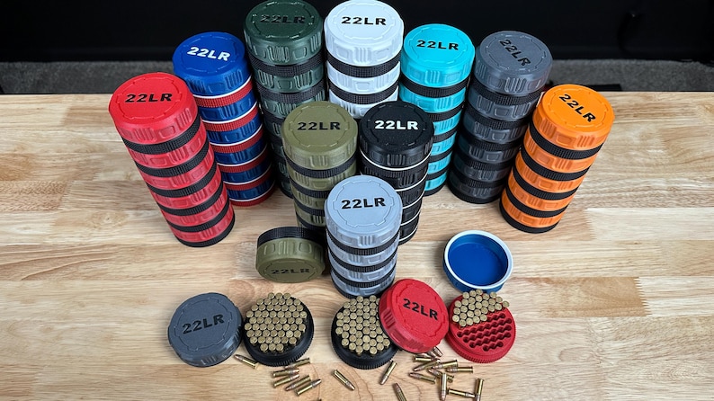 May include: A collection of cylindrical 22LR ammunition storage containers in various colors including red, blue, green, white, turquoise, grey, black, and orange. Each container features a textured grip and a lid marked with 22LR text.