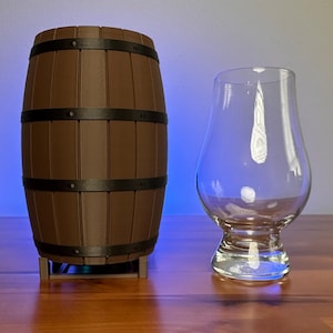 Puede incluir: Un barril de madera marrón con aros de metal se encuentra sobre una superficie de madera junto a un vaso de vidrio transparente.