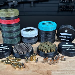 May include: A selection of ammunition containers with coloured lids, stacked and arranged on a wooden surface. The containers are black with coloured lids and feature the text "COME AND TAKE IT" along with calibre designations like "9mm" and "45acp".