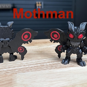 Puede incluir: Dos figuras de Mothman con cuerpos negros, alas texturizadas, patrones circulares rojos en las alas y ojos rojos brillantes. La palabra Mothman est&aacute; impresa en rojo sobre ellas. Las figuras tienen orejas puntiagudas y pies con garras, mostradas de frente y de espaldas.