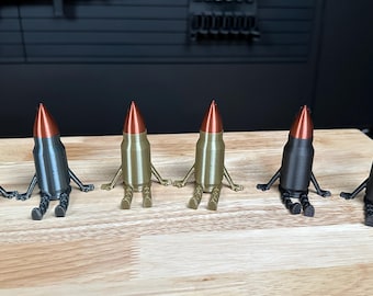 May include: Six small figurines shaped like bullets, each featuring a copper-colored tip and a cylindrical body with attached arms and legs. Four figurines are matte black, and two are metallic gold, all posed in a seated position.