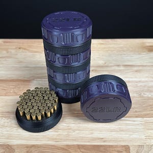 May include: A cylindrical purple and black storage container with a textured grip, featuring the text 22LR embossed on the lid. The container is stacked in sections, with one base section detached and filled with numerous small brass-colored 22LR cartridges.