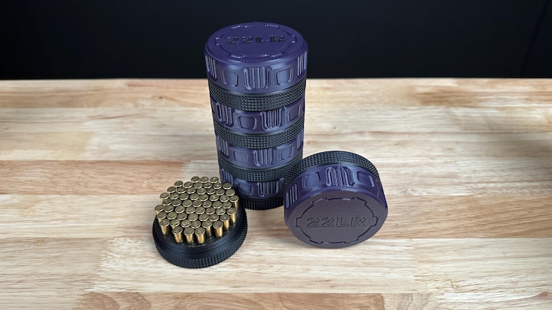 May include: A cylindrical purple and black storage container with a textured grip, featuring the text 22LR embossed on the lid. The container is stacked in sections, with one base section detached and filled with numerous small brass-colored 22LR cartridges.