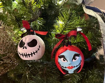 May include: Two Christmas ornaments featuring characters from The Nightmare Before Christmas. One is white with a black face, the other is red and blue. Both have red ribbons and hang on a Christmas tree.