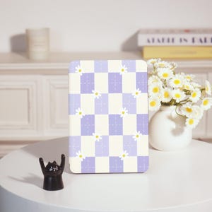 May include: A rectangular notebook with a checkerboard pattern in lavender and cream, adorned with small daisy illustrations. A black cat figurine with raised arms sits on a white table. A vase of white flowers with yellow centers is also on the table.