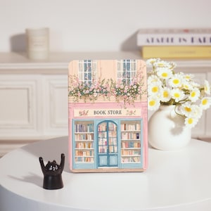 May include: A decorative wooden book-shaped sign depicting a bookstore facade. The sign is painted in pink and blue, with the words "BOOK STORE" at the top. A small black cat ornament is in front of the sign. The sign is on a white table.