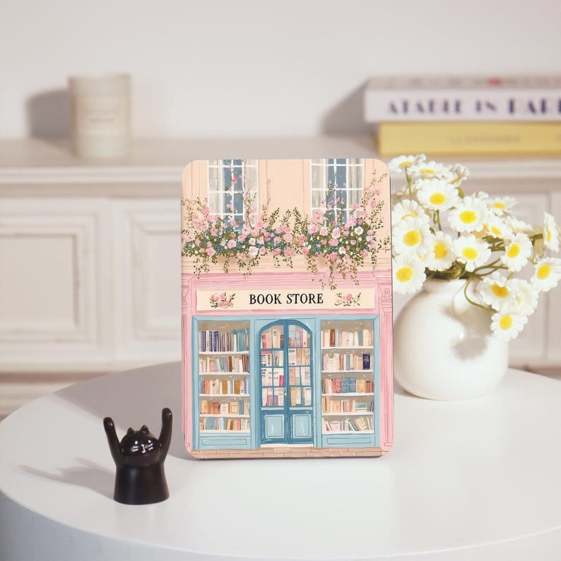 Op de afbeelding: Decoratief houten bord in de vorm van een boek met een illustratie van een boekhandelfa&ccedil;ade. Het bord heeft een roze en blauw kleurenschema, met de tekst "BOOK STORE" bovenaan. Een klein zwart kattenbeeldje staat ervoor.