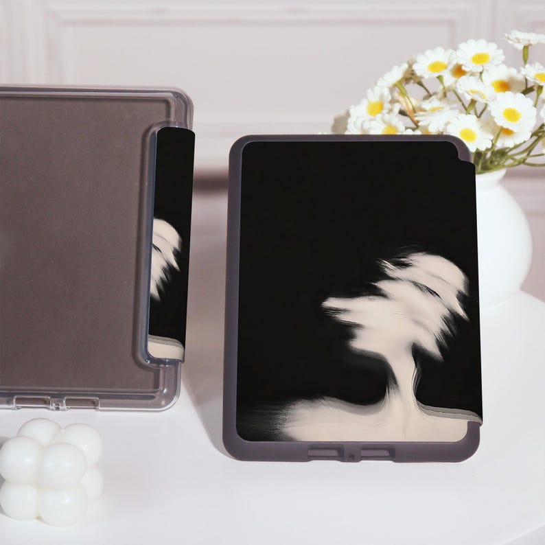 Puede incluir: Una funda negra para tableta con un dise&ntilde;o abstracto blanco. La funda est&aacute; al lado de una funda transparente para tableta. Un jarr&oacute;n con flores blancas y amarillas est&aacute; en el fondo.