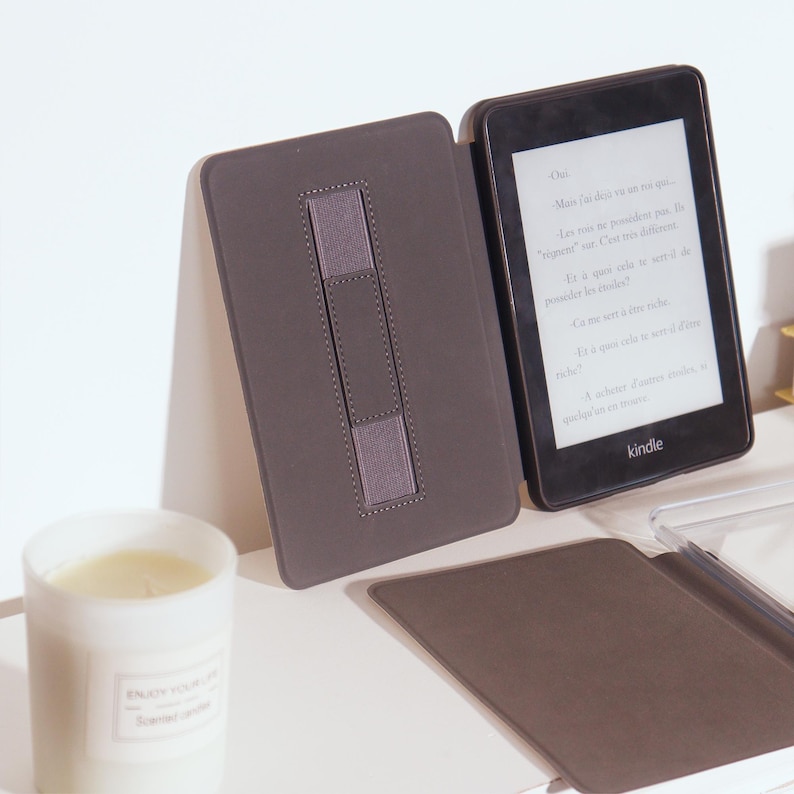 Op de afbeelding: Een Kindle e-reader met een donkergrijze hoes en een bijpassende standaard. De e-reader geeft tekst weer op het scherm. Een witte kaars met de tekst "ENJOY YOUR LIFE" staat op de voorgrond.