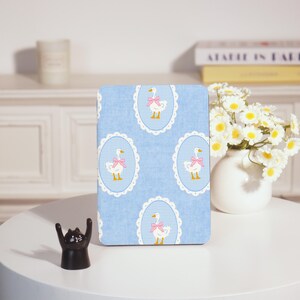 May include: A rectangular, light blue decorative item with a repeating pattern of white geese wearing pink bows inside oval frames. The background has a gingham pattern. The item is on a white table.