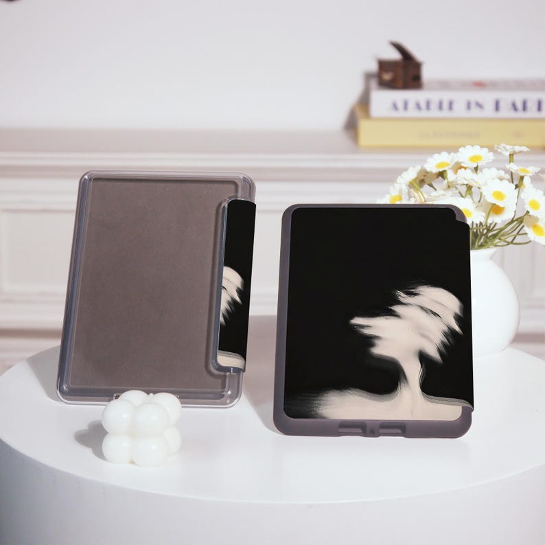 Puede incluir: Dos fundas para tabletas se muestran sobre una superficie blanca. Una es transparente y la otra es negra con un dise&ntilde;o abstracto blanco. Una peque&ntilde;a vela blanca con forma de burbuja se encuentra frente a las fundas. Un jarr&oacute;n con flores blancas est&aacute; en el fondo.