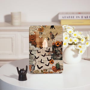 Könnte beinhalten: Eine Tablet-Hülle zum Thema Halloween mit einer Collage aus gruseligen Bildern. Das Design umfasst Kürbisse, Fledermäuse, Geister, Skelette und Hexen. Textfragmente lauten "witches go riding" und "Halloween". Die Hülle wird auf einem weißen Tisch mit einer schwarzen Katzenfigur präsentiert.