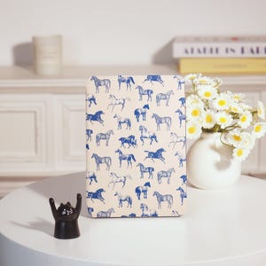 Peut inclure: Un carnet rectangulaire avec un motif répété de chevaux bleus sur fond crème. Le carnet est posé sur une table blanche. Une figurine de chat noir est visible au premier plan, ainsi que des fleurs.