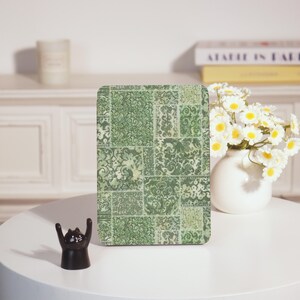 May include: A rectangular object with a green patchwork design, featuring floral and scroll patterns. It sits on a white table, next to a black cat figurine with raised arms and a vase of white flowers.