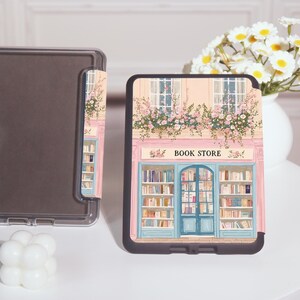 Op de afbeelding: Een zwarte tablethoes met een gedetailleerde illustratie van een boekhandelfa&ccedil;ade. Het ontwerp omvat een blauwe deur, boekenplanken vol boeken en de tekst "BOOK STORE". De hoes is versierd met roze bloemen.