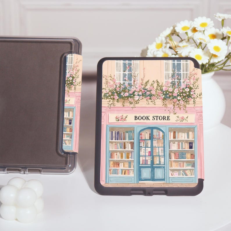 Op de afbeelding: Een zwarte tablethoes met een gedetailleerde illustratie van een boekhandelfa&ccedil;ade. Het ontwerp omvat een blauwe deur, boekenplanken vol boeken en de tekst "BOOK STORE". De hoes is versierd met roze bloemen.