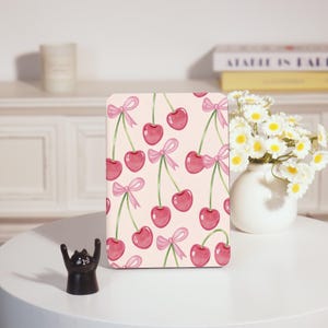 May include: A rectangular item with a pink background, adorned with a repeating pattern of red cherries and pink bows. The cherries and bows are painted in watercolour style. The item is displayed on a white surface, with a black cat figurine and a vase of flowers nearby.