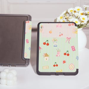 Peut inclure: Une coque rayée noire et blanche avec un motif rose et vert représentant des cerises, des fleurs et d'autres petits détails. La coque est conçue pour une liseuse électronique.