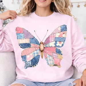 Puede incluir: Sudadera rosa claro con un diseño de mariposa grande. Las alas de la mariposa están hechas de retazos de mezclilla y tela en tonos azules, rosas y morados. La mariposa tiene antenas marrones y un cuerpo beige.