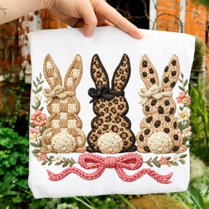 Puede incluir: Bolso tote blanco con tres conejos bordados con diseños variados. Los conejos presentan patrones a cuadros, leopardo y lunares, cada uno adornado con un lazo. Un lazo rosa tejido y detalles florales completan el diseño, con temática de Pascua.