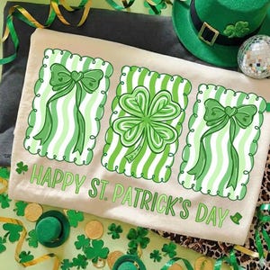 Puede incluir: Una camiseta color crema con un diseño del Día de San Patricio. La camiseta presenta tres rectángulos a rayas verdes y blancas con un lazo, un trébol de cuatro hojas y un diseño de lazo. Las palabras "HAPPY ST. PATRICK'S DAY" están impresas debajo.