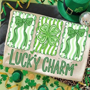 Puede incluir: Camiseta beige con un diseño de rayas verdes y blancas con un trébol de cuatro hojas y lazos. El texto "LUCKY CHARM" está impreso en verde. La imagen incluye decoraciones temáticas del Día de San Patricio.
